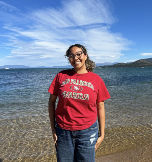 Jada Brooks is a programmer at the Women's Center. She is smiling in front a coastal view, and is wearing a red San Francisco 49ers shirt. 