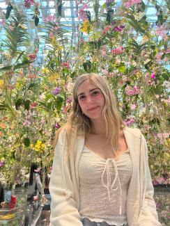 Kailey in a white shirt and cardigan in front of a many green, pink, and yellow flowers